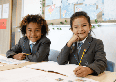 Two pupils from Cheadle Hulme Primary School smile as they sit at a desk doing work.